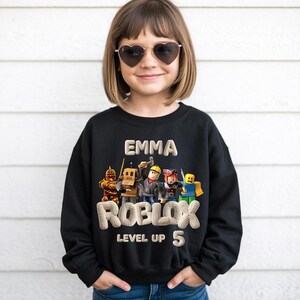 Puede incluir: Sudadera negra con el nombre "EMMA" y la palabra "ROBLOX" con "LEVEL UP 5" debajo. El dise&ntilde;o incluye personajes coloridos de Roblox. La ni&ntilde;a lleva gafas de sol en forma de coraz&oacute;n.