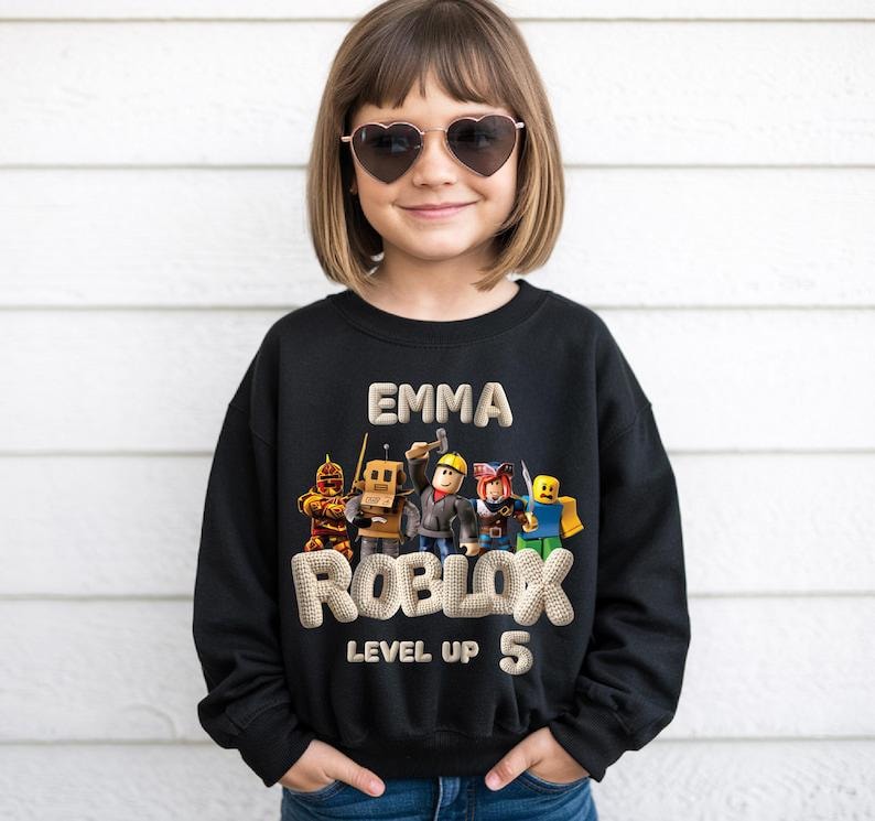 Puede incluir: Sudadera negra con el nombre "EMMA" y la palabra "ROBLOX" con "LEVEL UP 5" debajo. El dise&ntilde;o incluye personajes coloridos de Roblox. La ni&ntilde;a lleva gafas de sol en forma de coraz&oacute;n.