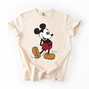 May include: Cream-colored t-shirt featuring a classic Mickey Mouse graphic. The cartoon character is depicted in his iconic pose, wearing red shorts, brown shoes, and a black top. The Disney logo is visible.