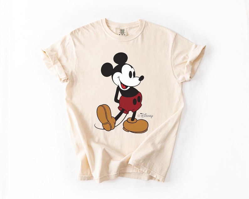May include: Cream-colored t-shirt featuring a classic Mickey Mouse graphic. The cartoon character is depicted in his iconic pose, wearing red shorts, brown shoes, and a black top. The Disney logo is visible.
