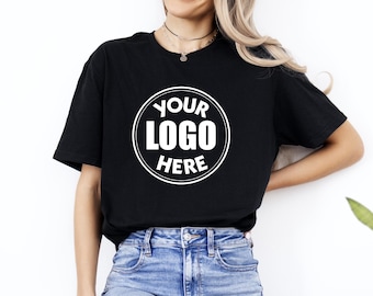 May include: Black t-shirt with a white circular logo that reads "YOUR LOGO HERE". The shirt is a classic crew neck style and is paired with blue denim jeans. The model is wearing a necklace.