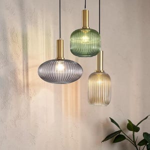 May include: Three pendant lights with ribbed glass shades in green, gray, and amber. Each light has a gold-colored metal fixture and a black cord. The lights are hanging against a neutral-colored wall.