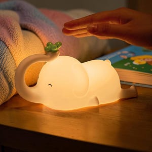 May include: A soft, glowing white elephant-shaped night light with a green clover on its trunk. The light is on a wooden surface, with a hand hovering above it. A colorful blanket and a book are in the background.