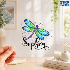 May include: A clear acrylic plaque featuring a colorful dragonfly with blue and green wings and a purple body. Below the dragonfly, the name Sophia is printed in a black cursive script font with small heart accents. The design includes small stars.