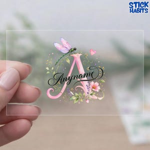 May include: A clear adhesive sticker featuring a large pink letter A decorated with watercolor-style flowers, green leaves, and a dragonfly with purple wings. The word Anyname is written in black cursive script across the center of the letter.