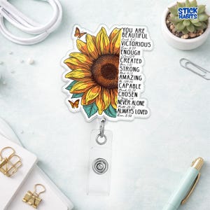 May include: A badge reel featuring a vibrant sunflower design with orange and yellow petals, green leaves, and two butterflies. The badge reel has the words "YOU ARE BEAUTIFUL" and other inspirational words. The reel has a clear plastic holder.