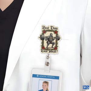Op de afbeelding: Een rechthoekige badge reel met een ridder die gewichten heft, omringd door rozen. De tekst "Dost Thou Even Hoist?" staat in een decoratief lettertype. De badge reel is bevestigd aan een witte labjas.