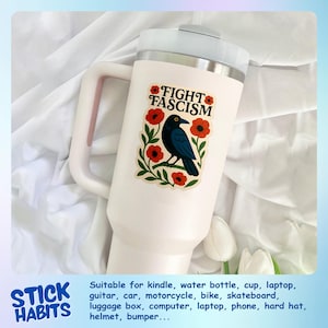 May include: A white insulated tumbler with a handle, featuring a sticker that reads "FIGHT FASCISM." The sticker showcases a black bird, red flowers, and green leaves. The tumbler is designed for cold beverages.