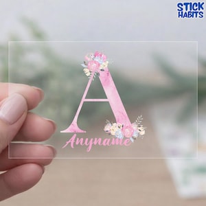 May include: A clear rectangular sticker featuring a large pink watercolor letter A decorated with floral illustrations in shades of pink, blue, and cream. Below the letter, the word Anyname is printed in a cursive pink script font.