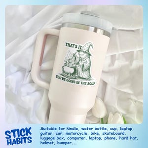 May include: A large, off-white tumbler with a handle and a silver lid. A green and white sticker on the tumbler reads "THAT'S IT, YOU'RE GOING IN THE SOUP" with an illustration of a frog in a wizard hat. The tumbler is suitable for water bottles and laptops.