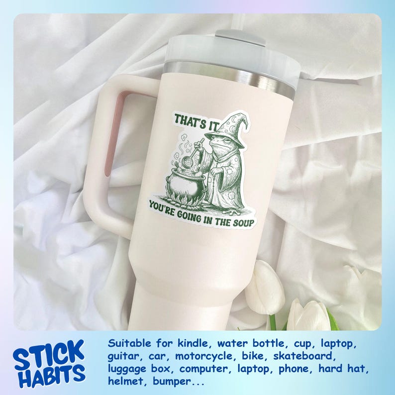 May include: A large, off-white tumbler with a handle and a silver lid. A green and white sticker on the tumbler reads "THAT'S IT, YOU'RE GOING IN THE SOUP" with an illustration of a frog in a wizard hat. The tumbler is suitable for water bottles and laptops.
