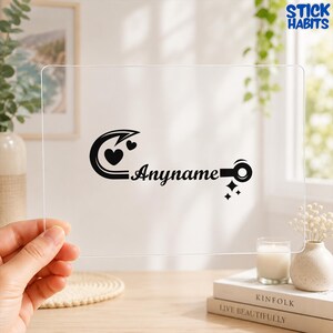 May include: A clear rectangular acrylic sheet featuring a black vinyl decal. The design includes a stylized fishing hook shaped like a heart, the word Anyname in a cursive script, and a circular end with small star accents near the bottom.