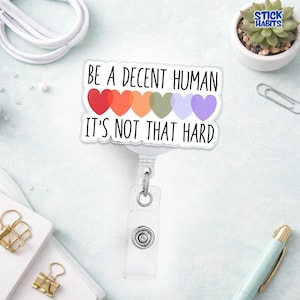 May include: A white retractable badge reel with the text "BE A DECENT HUMAN IT'S NOT THAT HARD" above a row of colorful hearts. The hearts are red, orange, yellow, green, light blue, and purple. The reel has a clear plastic strap and a metal clip.