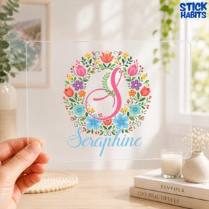 Peut inclure: Une plaque en acrylique transparent ornée d'une grande lettre S rose en cursive au centre d'une couronne florale colorée composée de fleurs rouges, roses, violettes, orange et bleues avec des feuilles vertes. Le nom Seraphine est écrit en bleu.