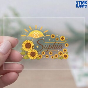 May include: A clear sticker featuring a sunflower design with the name "Sophia" in brown script. The design includes a yellow sun, sunflowers, and stars. The sticker is held by a hand.
