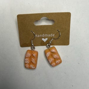 May include: A pair of dangle earrings featuring rectangular charms shaped like braided bread loaves. The charms are orange with white highlights to mimic a baked crust texture. They hang from silver-toned metal hooks attached to a brown display card.