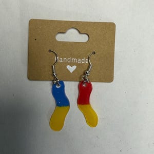 May include: A pair of dangle earrings featuring wavy, translucent acrylic charms attached to silver-toned metal hooks. One charm is blue and yellow, while the other is red and yellow. The earrings are displayed on a brown cardboard tag with the text Handmade and a small white heart icon.