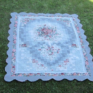 May include: A gray and white quilt with a scalloped edge and a floral pattern. The quilt is made of squares and has a light blue background.