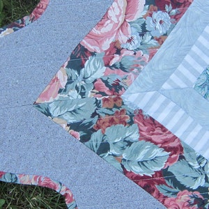 May include: A close-up of a quilt with a floral pattern in shades of pink, green, and white. The quilt is made of different fabrics, including a solid gray fabric and a striped fabric.
