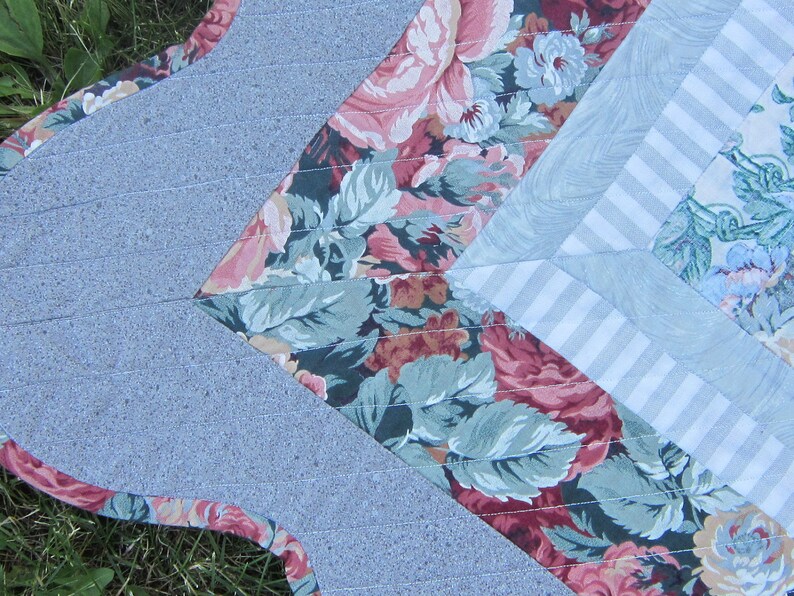 May include: A close-up of a quilt with a floral pattern in shades of pink, green, and white. The quilt is made of different fabrics, including a solid gray fabric and a striped fabric.