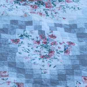 May include: A patchwork quilt with a floral pattern in shades of pink and white. The quilt is made of squares of different fabrics, and the floral pattern is repeated throughout. The quilt is folded in half, and the fabric is slightly wrinkled.