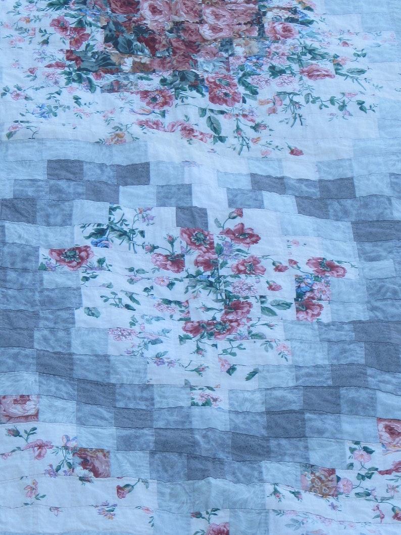 May include: A patchwork quilt with a floral pattern in shades of pink and white. The quilt is made of squares of different fabrics, and the floral pattern is repeated throughout. The quilt is folded in half, and the fabric is slightly wrinkled.