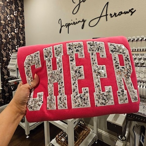 May include: A bright pink sweatshirt featuring the word CHEER in large, block-style letters. Each letter is filled with shimmering silver sequins and outlined with a white border, creating a bold, textured appearance against the solid pink fabric.