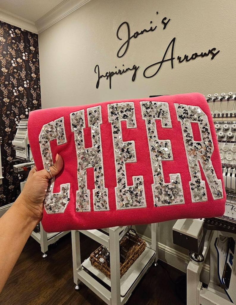 May include: A bright pink sweatshirt featuring the word CHEER in large, block-style letters. Each letter is filled with shimmering silver sequins and outlined with a white border, creating a bold, textured appearance against the solid pink fabric.