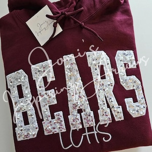 May include: A maroon hooded sweatshirt featuring the word BEARS in large, white-bordered block letters filled with silver sequins. A white tag with the text Joni's Inspiring Arrows is attached to the drawstring, and the letters LCHS appear below.