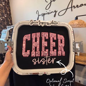 May include: A white embroidery hoop holding black fabric with the word CHEER in large pink sequined block letters and the word sister in pink script lettering. The hoop features the text Joni's Inspiring Arrows at the top center.
