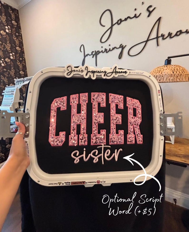 May include: A white embroidery hoop holding black fabric with the word CHEER in large pink sequined block letters and the word sister in pink script lettering. The hoop features the text Joni's Inspiring Arrows at the top center.
