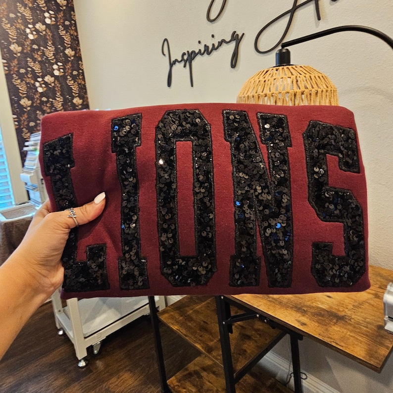 May include: A maroon sweatshirt featuring the word LIONS in large, black, sequined block letters. The sequins are densely applied to the fabric, creating a textured and reflective surface across each letter of the text displayed on the front.