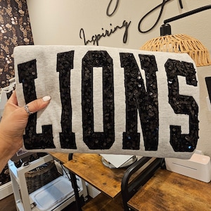 May include: A light gray sweatshirt featuring the word LIONS in large, bold, black sequined block letters across the front. The sequins are densely packed, creating a textured, reflective surface that contrasts with the smooth, heathered fabric.