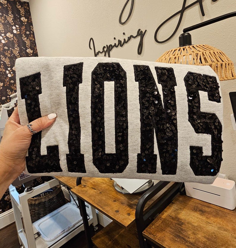 May include: A light gray sweatshirt featuring the word LIONS in large, bold, black sequined block letters across the front. The sequins are densely packed, creating a textured, reflective surface that contrasts with the smooth, heathered fabric.