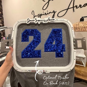 May include: A white rectangular embroidery hoop holding a grey fabric garment. The fabric features the number 24 in large, bright blue sequins with a dark blue border. A white sign with black text reads Joni's Inspiring Arrows at the top.