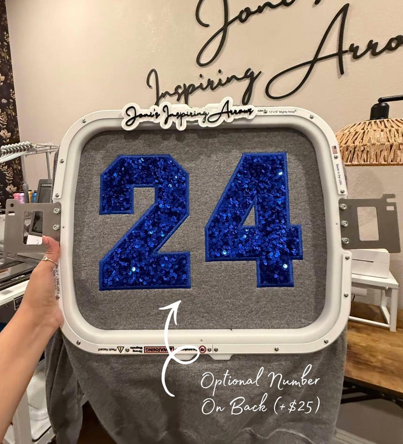 May include: A white rectangular embroidery hoop holding a grey fabric garment. The fabric features the number 24 in large, bright blue sequins with a dark blue border. A white sign with black text reads Joni's Inspiring Arrows at the top.