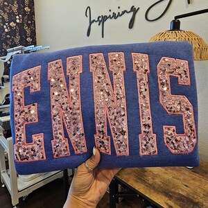 May include: A blue sweatshirt featuring the word ENNIS in large, block-style letters. The letters are filled with shimmering, light pink sequins and outlined with a matching pink fabric border, creating a textured and reflective appearance.