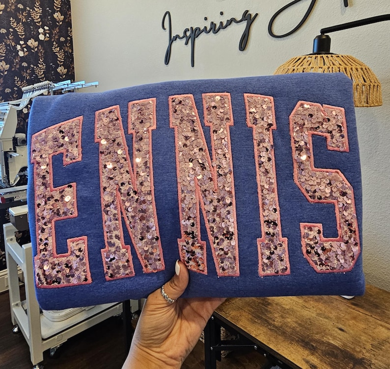 May include: A blue sweatshirt featuring the word ENNIS in large, block-style letters. The letters are filled with shimmering, light pink sequins and outlined with a matching pink fabric border, creating a textured and reflective appearance.
