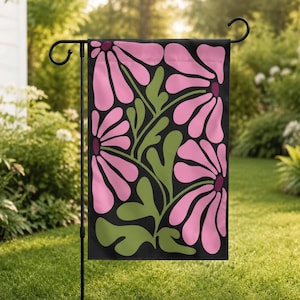 May include: A garden flag featuring a bold floral design. The flag displays large pink flowers with dark purple centers and green leaves against a black background. The flag is hanging on a black metal stand in a grassy yard.
