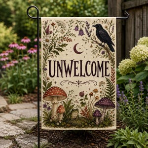 May include: A rectangular garden flag featuring the text UNWELCOME in dark lettering. The design includes a black crow perched on a branch, a crescent moon, various wildflowers, ferns, and several mushrooms with a small frog sitting at the base.