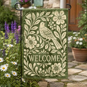 Green welcome garden flag with bird and floral folk art design displayed on a black flag stand in a garden