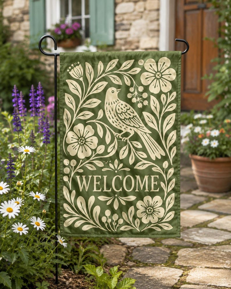 Green welcome garden flag with bird and floral folk art design displayed on a black flag stand in a garden