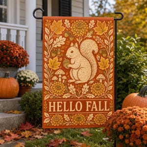 Pode incluir: Uma bandeira de jardim retangular com fundo laranja apresentando uma ilustração central de um esquilo segurando uma bolota. O design inclui folhas de carvalho, bolotas, cogumelos e crisântemos. O texto HELLO FALL está impresso em branco.