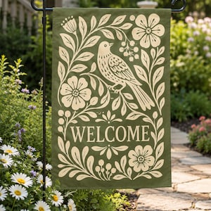 May include: A rectangular garden flag in olive green featuring a cream-colored screen-printed design of a bird perched on a branch surrounded by flowers and foliage. The word WELCOME is printed in a serif font in the center of the flag.