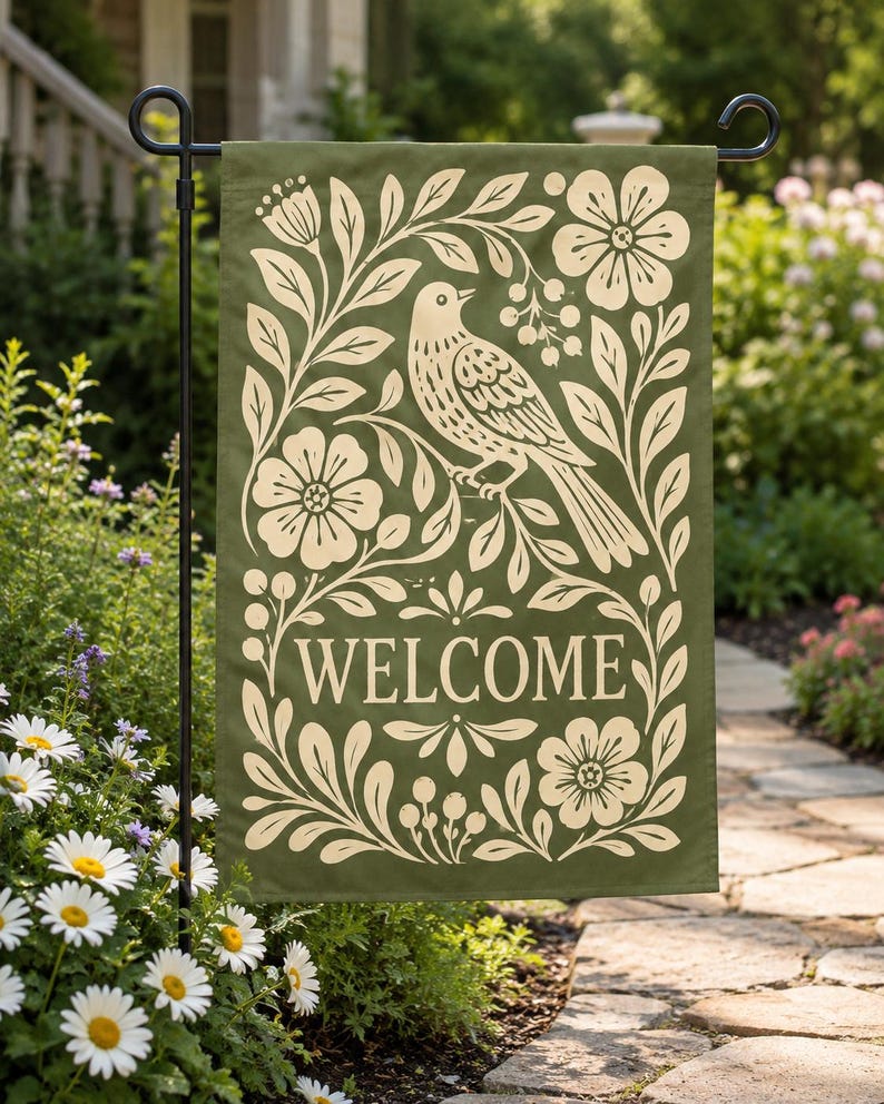 Green welcome garden flag with bird and floral folk art design displayed on a black flag stand in a garden