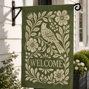 May include: A rectangular garden flag in olive green featuring a cream-colored graphic print of a bird perched on a branch surrounded by floral motifs and leaves. The word WELCOME is printed in a serif font in the lower center of the flag.