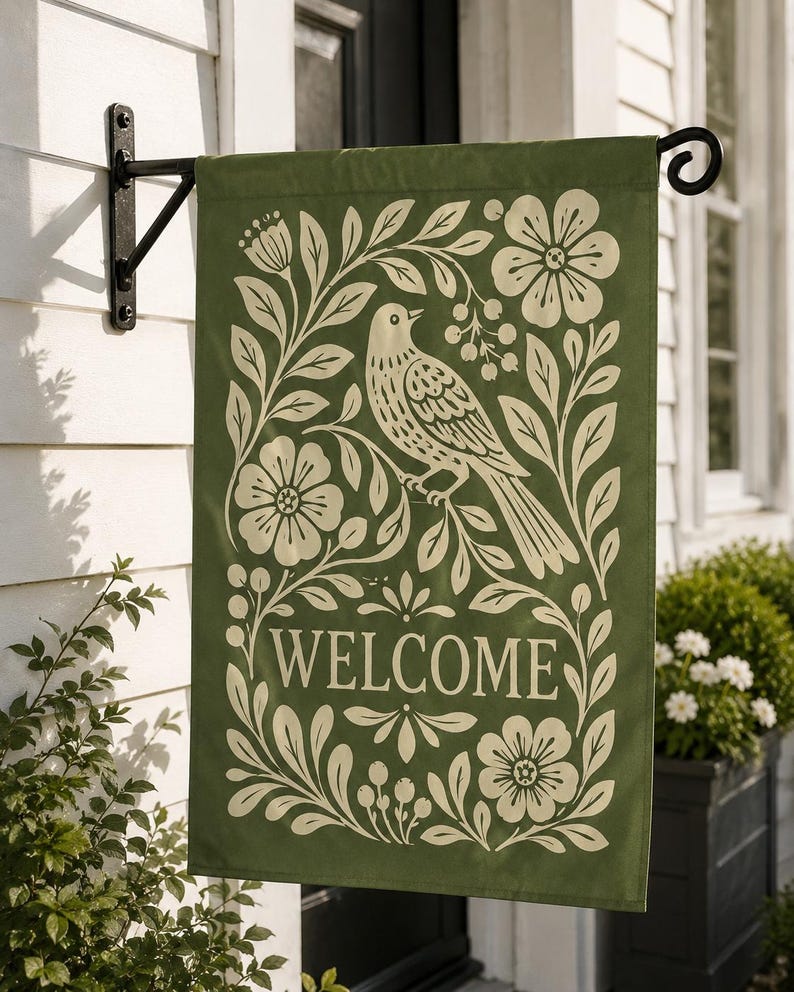 May include: A rectangular garden flag in olive green featuring a cream-colored graphic print of a bird perched on a branch surrounded by floral motifs and leaves. The word WELCOME is printed in a serif font in the lower center of the flag.
