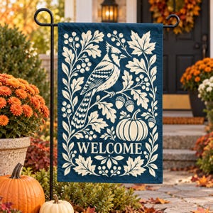 Puede incluir: Una bandera de jardín rectangular en azul oscuro con una ilustración color crema de un cardenal posado en una rama, rodeado de hojas de roble, bayas y bellotas. Una calabaza se sitúa bajo el ave y la palabra WELCOME está impresa.