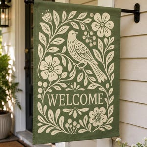 May include: A rectangular garden flag in olive green featuring a cream-colored graphic print of a bird perched on a branch surrounded by flowers, leaves, and berries. The word WELCOME is printed in a bold, serif font at the bottom center.