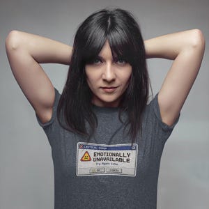 Puede incluir: Una camiseta gris oscuro con el texto "EMOTIONALLY UNAVAILABLE" en el estilo de un mensaje de error de computadora. La camiseta presenta un icono de cara triste y las palabras "Try Again Later". La persona de la imagen tiene cabello oscuro.